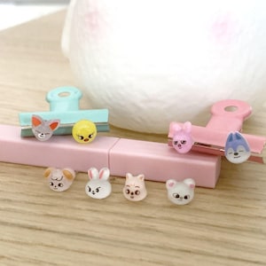 Può includere: Una collezione di piccole clip e ciondoli decorativi con facce di animali. Le clip sono azzurre e rosa, con facce di animali. I ciondoli includono un pulcino giallo, una volpe grigia, un coniglio rosa e un lupo blu.