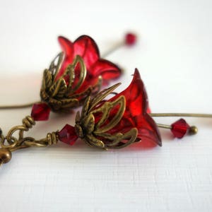 May include: A pair of dangle earrings featuring translucent red bell-shaped flower beads encased in ornate antique brass-toned metal caps. Each earring includes small faceted red crystal beads and thin metal stems ending in tiny red bead accents.
