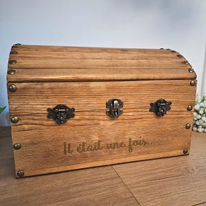 May include: A wooden chest with a curved top, metal hardware, and the phrase "Il était une fois..." engraved on the front. The chest is made of light brown wood and has a vintage aesthetic.