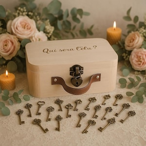 Può includere: Una scatola di legno chiaro con manico e serratura a forma di cuore, circondata da chiavi antiche. La scatola ha il testo "Qui sera l'élu?" inciso. La scena comprende candele e rose.