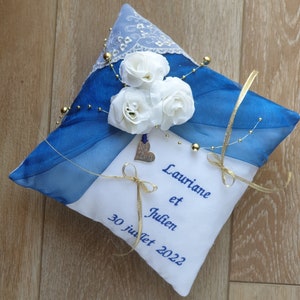 May include: A white ring pillow with blue and white satin fabric, decorated with white flowers and gold beads. The pillow is embroidered with the names "Lauriane et Julien" and the date "30 juillet 2022".