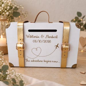 Puede incluir: Una caja de tarjetas rectangular blanca con forma de maleta vintage, asa superior, correas metálicas doradas y esquinas reforzadas. Incluye el texto "Wiktoria & Norbert", "08/10/2026" y "The adventure begins now..." con un gráfico de corazón y avión.