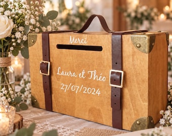 May include: A rectangular wooden box designed like a vintage suitcase with a top handle, dark brown leather straps, and brass-toned corner accents. The front features a horizontal slot and white text reading Merci, Laura et Théo, 27/07/2024.