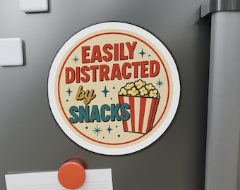 May include: A round magnet with the text "EASILY DISTRACTED by SNACKS" in red and blue. A popcorn bucket graphic is on the right. The magnet is on a gray surface with other magnets.