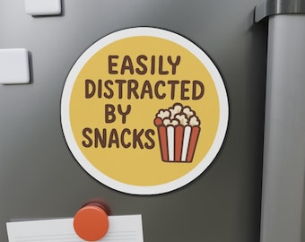 May include: A round magnet with the text "EASILY DISTRACTED BY SNACKS" in brown letters on a yellow background. A cartoon popcorn bucket is below the text. The magnet is on a gray surface.