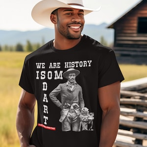 May include: A black t-shirt with a white graphic of a man in a cowboy hat and the text "WE ARE HISTORY ISOM DART THAT MELANIN THO".