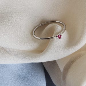 May include: A silver ring with a small, heart-shaped, red gemstone. The ring is set against a white fabric background, with a hint of blue fabric in the lower portion of the image. The ring's band is thin and delicate.