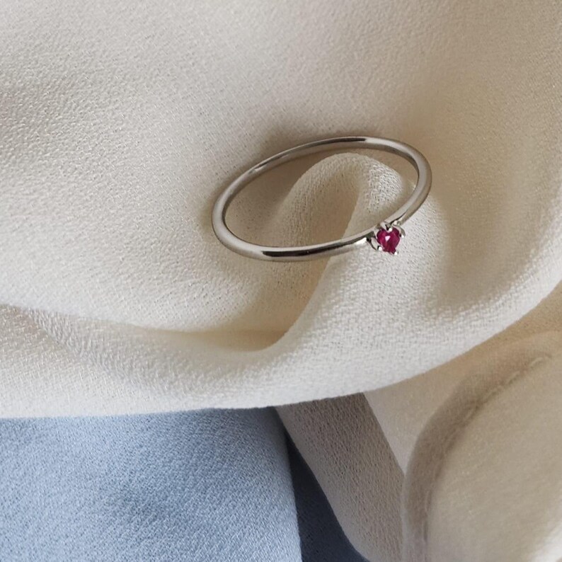 May include: A silver ring with a small, heart-shaped, red gemstone. The ring is set against a white fabric background, with a hint of blue fabric in the lower portion of the image. The ring's band is thin and delicate.