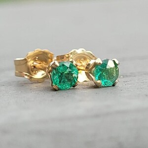 May include: A pair of gold stud earrings featuring round, vivid green gemstones. The earrings are set in a classic four-prong setting, highlighting the bright green colour of the stones. The gold setting and posts are visible.