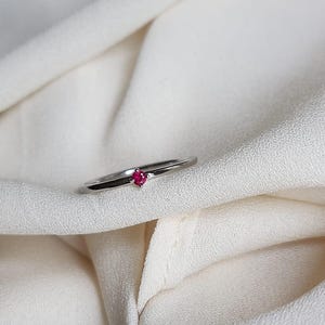 May include: A silver ring with a small, round, bright red gemstone set in the center. The ring is displayed on a white, textured fabric. The ring is a piece of jewelry.