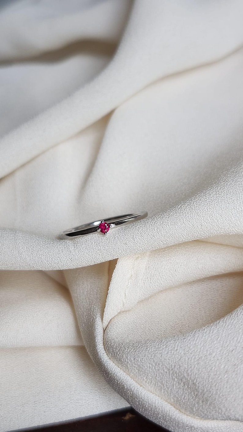 May include: A silver ring with a small, round, bright red gemstone set in the center. The ring is displayed on a white, textured fabric. The ring is a piece of jewelry.