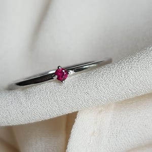 May include: A delicate silver ring featuring a small, vibrant red gemstone. The ring's slender band and the bright red stone create a minimalist and elegant design. The ring is set against a soft, white fabric background.