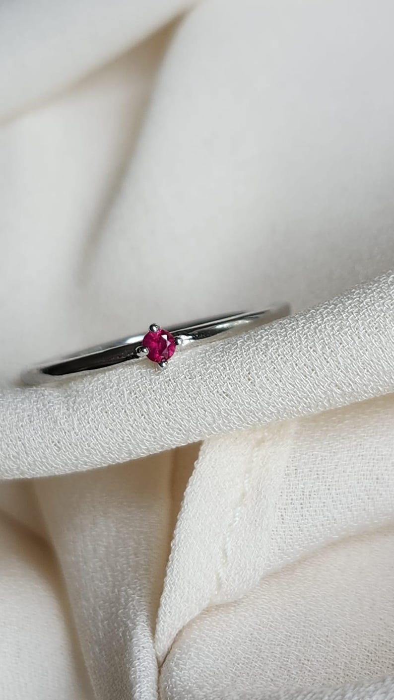 May include: A delicate silver ring featuring a small, vibrant red gemstone. The ring's slender band and the bright red stone create a minimalist and elegant design. The ring is set against a soft, white fabric background.