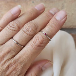 May include: Two delicate silver rings are displayed on a hand. One ring features a small, clear gemstone, while the other has a tiny red gemstone. The rings are simple and elegant, suitable for everyday wear.