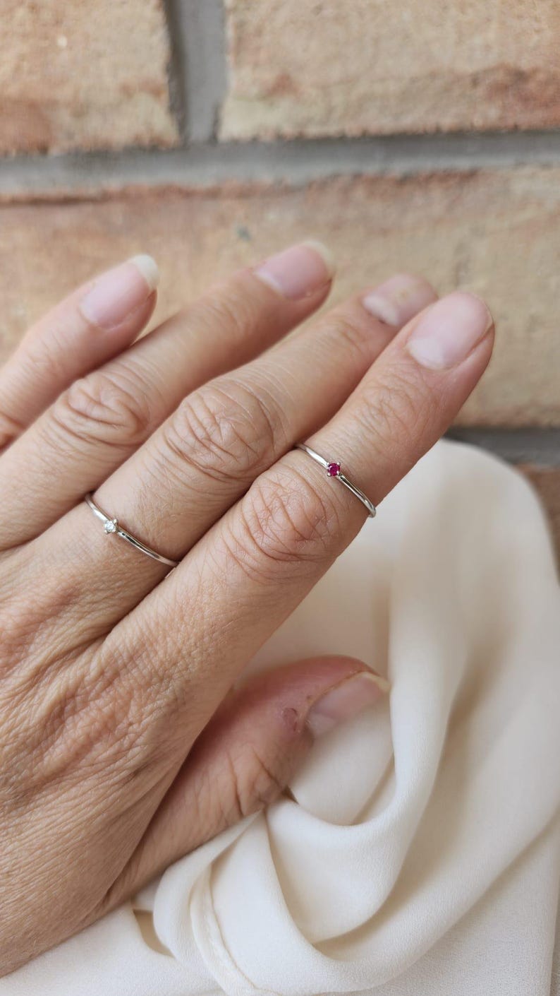 May include: Two delicate silver rings are displayed on a hand. One ring features a small, clear gemstone, while the other has a tiny red gemstone. The rings are simple and elegant, suitable for everyday wear.