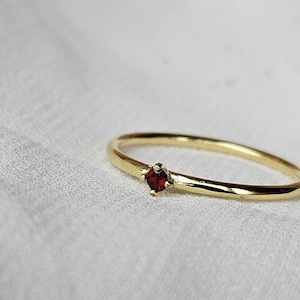 May include: A delicate gold band ring with a single, small, red gemstone set in the centre.