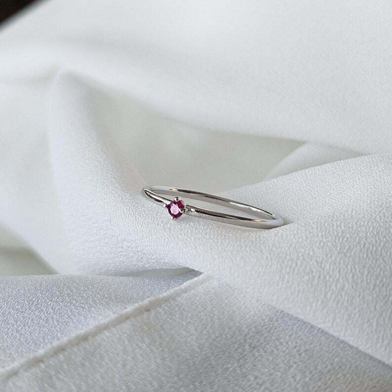 May include: A delicate silver ring with a small, round, ruby-colored gemstone set in the center. The ring is displayed on a white, textured fabric, creating a minimalist and elegant aesthetic. The band is thin and polished, highlighting the gemstone.