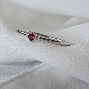 May include: A delicate silver ring featuring a small, round, deep red gemstone. The ring has a thin band and a simple, elegant design. The gemstone is set in a classic four-prong setting, adding a touch of sparkle.