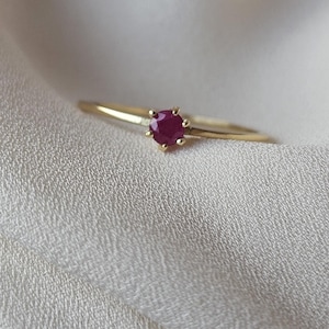 Peut inclure: Une fine bague en or ornée d'un rubis rond et rouge foncé. Le rubis est serti dans une monture à six griffes, et le corps de la bague est fin et poli. La bague est présentée sur un tissu blanc cassé.