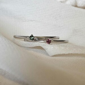 May include: Two delicate silver rings with small gemstones. The ring on the left has a green gemstone, and the ring on the right has a pink gemstone.