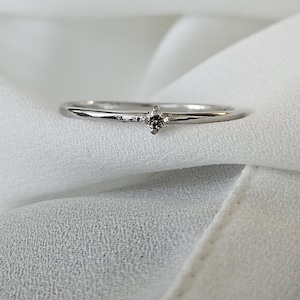 May include: A delicate silver ring featuring a small, round, light brown gemstone. The ring is set against a soft, white fabric background, accentuating its minimalist design and elegant simplicity. The band is thin and polished.