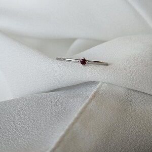 May include: A delicate silver ring featuring a small, round, deep red gemstone. The ring has a thin band and a minimalist design, set against a soft, white fabric background. The gemstone is the focal point.