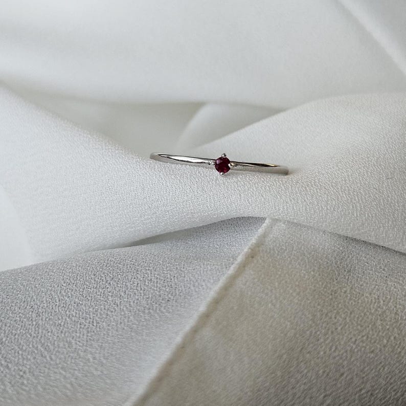 May include: A delicate silver ring featuring a small, round, deep red gemstone. The ring has a thin band and a minimalist design, set against a soft, white fabric background. The gemstone is the focal point.