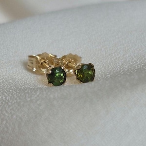 May include: A pair of stud earrings featuring round, faceted olive green gemstones set in gold metal prongs. The earrings include matching gold metal butterfly backings, showcasing a classic design with a polished finish and vibrant green hues.