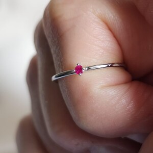 May include: A delicate silver ring featuring a small, round, bright red gemstone. The ring has a thin band and a minimalist design. The gemstone is set in a simple, four-prong setting, creating a classic and elegant look.