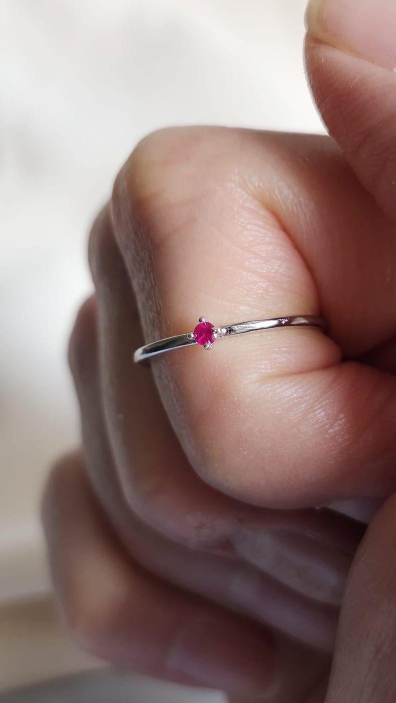 May include: A delicate silver ring featuring a small, round, bright red gemstone. The ring has a thin band and a minimalist design. The gemstone is set in a simple, four-prong setting, creating a classic and elegant look.