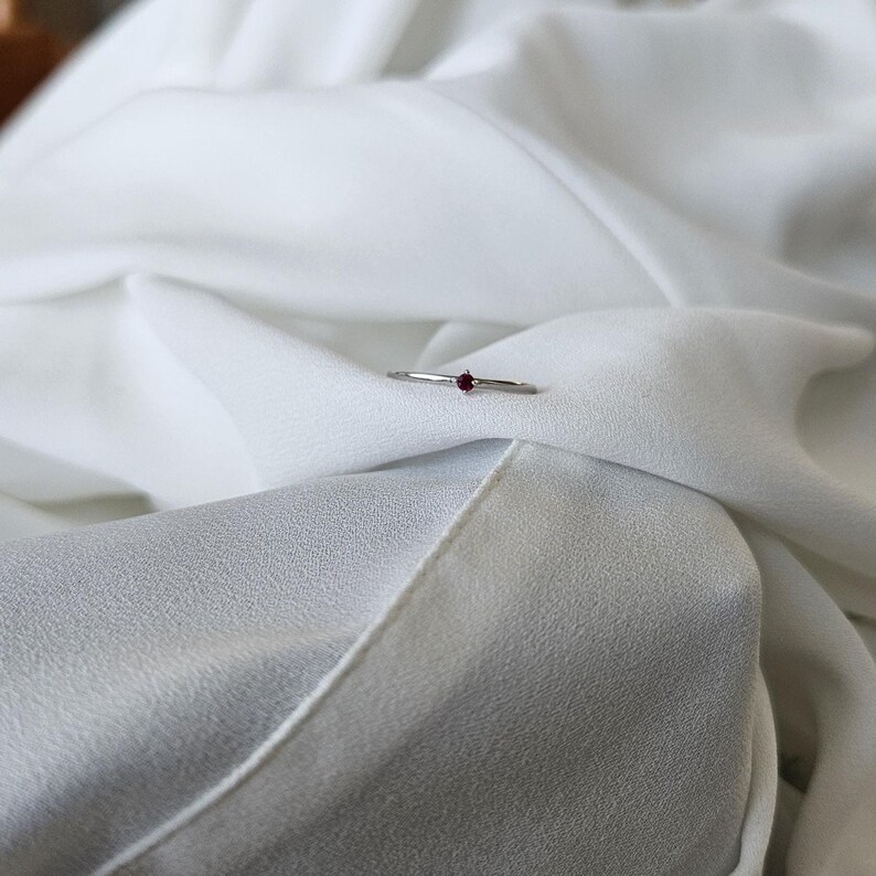May include: A delicate silver ring with a small, round, deep red gemstone set in the center. The ring is displayed on a white, flowing fabric, creating a minimalist and elegant aesthetic.