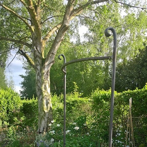 May include: A dark metal garden plant support with a rectangular frame and scrolled top. The support is set in a garden with a stone path, green grass, and various plants, including a large tree.