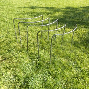 May include: Four metal plant supports with a curved top and straight legs are arranged on green grass. The supports are made of dark grey metal, designed to hold up plants. The supports are in a garden.