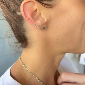 May include: A close-up of a person's ear with three gold earrings. The earrings are a small green stone, a small gold hexagon, and a longer gold bar with a turquoise stone. The earrings are connected by a delicate gold chain.