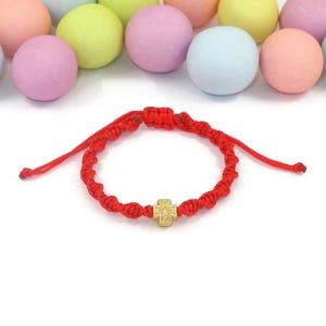 May include: A red cord bracelet featuring a small gold-toned cross charm with a central embossed star design. The bracelet is constructed with a series of intricate knots and includes adjustable pull strings. The text elizart shop is visible.