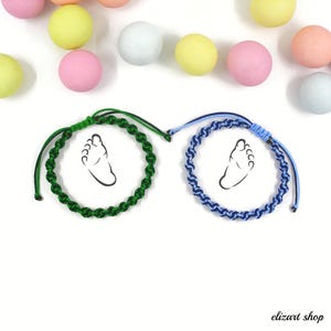 Puede incluir: Dos pulseras de macramé, una verde y otra azul, cada una con un diseño de huella negra. Las pulseras se exhiben sobre una superficie blanca con caramelos de colores esparcidos. Las pulseras son hechas a mano.