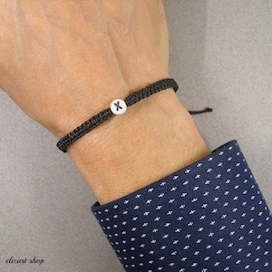 Könnte beinhalten: Schwarz geflochtenes Armband mit einer weißen Perle mit dem schwarzen Buchstaben "X". Das Armband wird am Handgelenk getragen, mit einem marineblauen Hemdsärmel mit Muster im Hintergrund. Schlichtes, minimalistisches Design.
