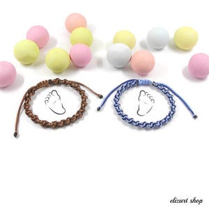 Könnte beinhalten: Zwei handgefertigte Armbänder mit einem Babyfußabdruck-Design. Eines ist braun, das andere blau und weiß. Pastellfarbene Kugeln in Rosa, Gelb und Weiß sind um die Armbänder herum verstreut. Die Armbänder sind wahrscheinlich für ein Baby oder einen neuen Elternteil.