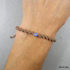 Puede incluir: Una pulsera de macramé marrón con un dije de ojo turco azul y blanco. La pulsera está anudada y es ajustable, con un diseño simple y minimalista. El dije es una pequeña cuenta redonda con un centro azul y detalles blancos y negros.