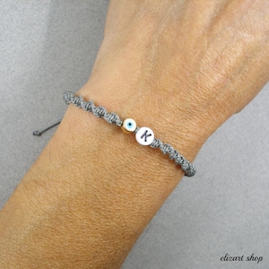 May include: Grey macrame bracelet featuring a gold bead, a white bead with the letter "K", and an evil eye charm. The adjustable bracelet is handmade. The bracelet is on a person's wrist.