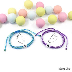 Puede incluir: Dos pulseras de amistad hechas a mano con un diseño de huella de bebé. Una pulsera es azul y verde, la otra es morada. Cada pulsera tiene una cuenta de letra blanca. Caramelos de colores pastel en el fondo.