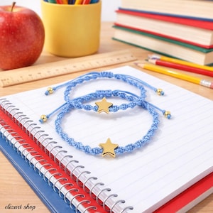 Puede incluir: Dos pulseras de macramé azul claro con dijes de estrella dorada se muestran en un cuaderno. Las pulseras están anudadas con cierres ajustables. Una manzana roja, lápices y libros están en el fondo. El texto "elizart shop" es visible.