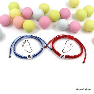 May include: Two handmade bracelets, one blue and one red, each featuring a baby footprint design and a single initial bead. The bracelets are displayed with colorful, round candies in shades of pink, yellow, and white.