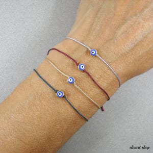 May include: Four delicate bracelets, each with a small, blue evil eye charm. The bracelets are strung on thin cords in various colours: dark teal, beige, burgundy, and grey. The charms have a blue centre with a white and black outline.