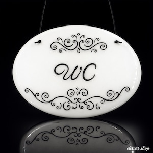May include: White oval sign with the letters "WC" in black script, framed by elegant black scrollwork. The sign is suspended by a black cord, and its reflection is visible below.