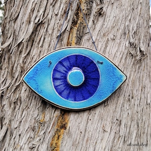 May include: A ceramic eye-shaped wall hanging in shades of blue. The eye has a textured surface and a dark blue centre, with a light blue outer edge. It is suspended by a blue string.