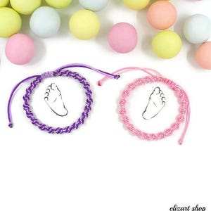 May include: Two circular macrame bracelets featuring adjustable cord closures. One bracelet is crafted from purple cord, and the other is made from light pink cord. Each bracelet centres a white circular charm displaying a black line drawing of a foot.