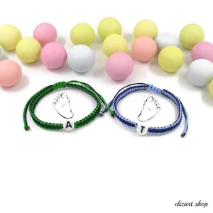 Peut inclure: Deux bracelets en macramé, un vert et un bleu, chacun avec une perle blanche portant une lettre. Les bracelets présentent un motif d'empreinte de bébé. Des bonbons aux couleurs pastel, roses, jaunes et blancs, sont dispersés en arrière-plan.