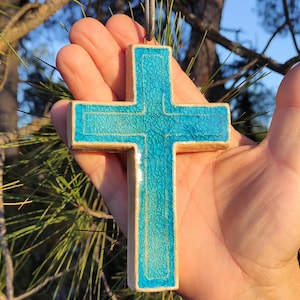 May include: A ceramic cross with a turquoise crackle glaze. The cross is held up by a metal wire. The cross has a light brown border. The background is blurred with green pine needles and sunlight.