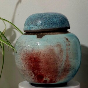 Op de afbeelding: Een keramische pot met een deksel, met een blauwe en bruine glazuur met een gekrakelde afwerking. De pot heeft een roodbruine vlek aan de zijkant.